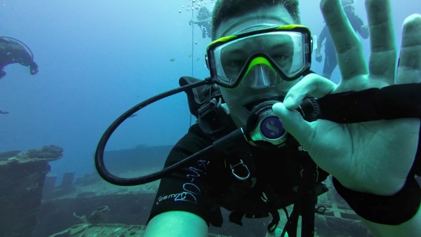 Photograph of Colton giving okay symbol with fingers while scuba diving above ship wreck.